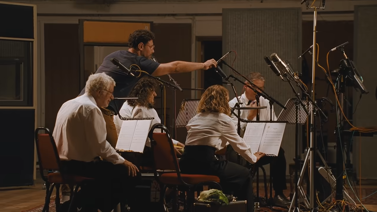 Members of the London Vegetable Orchestra sit with music stands and microphones in an Abbey Road recording studio while a sound engineer adjusts a microphone.