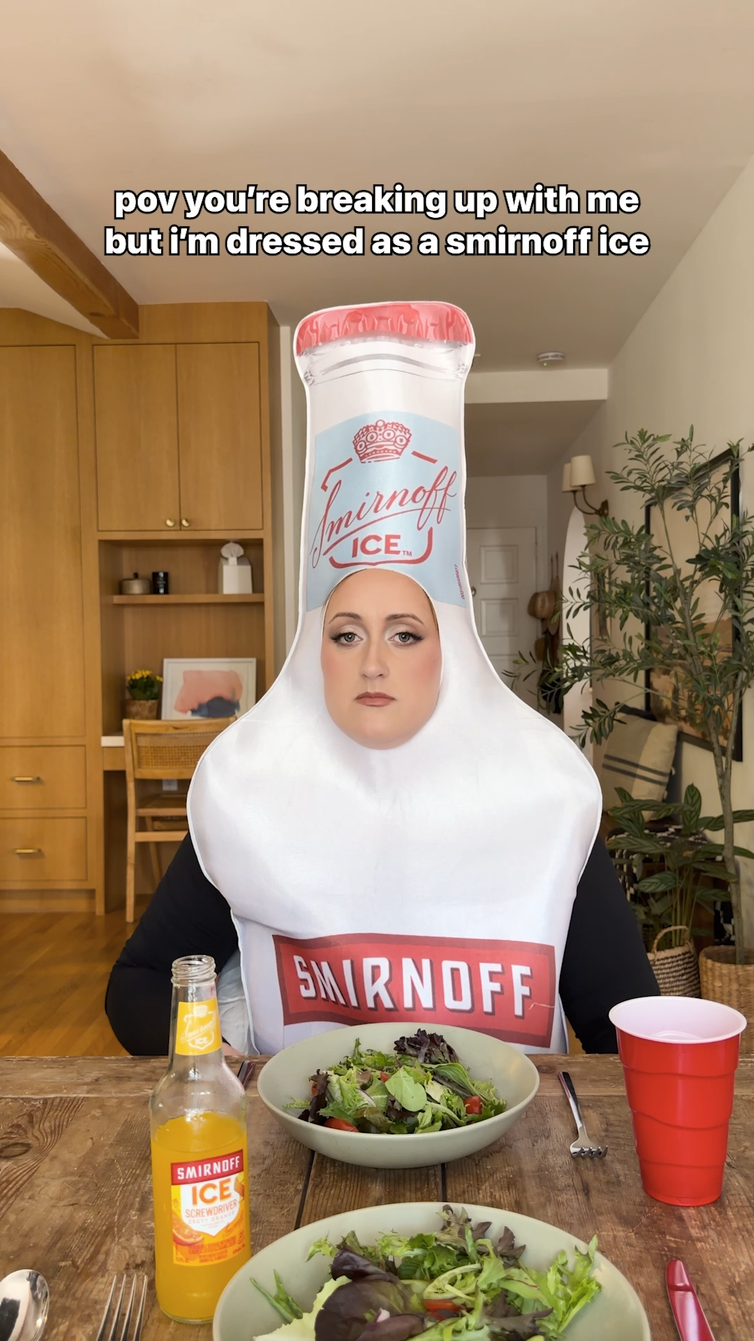 Brittany Broski sits at a table wearing an oversized Smirnoff Ice bottle costume beneath the caption “pov you’re breaking up with me but i’m dressed as a smirnoff ice”.